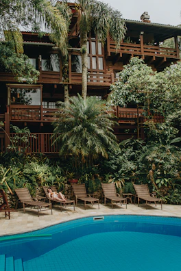 A lush tropical scene features a large wooden house surrounded by vibrant greenery, including palm trees and other lush plants. Below the house, a swimming pool reflects the blue sky. Several lounge chairs are placed along the poolside, and a person is relaxing on one of them, enjoying the tranquil atmosphere.