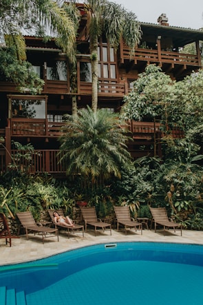 A lush tropical scene features a large wooden house surrounded by vibrant greenery, including palm trees and other lush plants. Below the house, a swimming pool reflects the blue sky. Several lounge chairs are placed along the poolside, and a person is relaxing on one of them, enjoying the tranquil atmosphere.