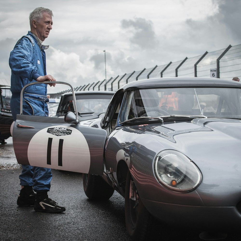 man in blue jacket standing beside silver volkswagen beetle