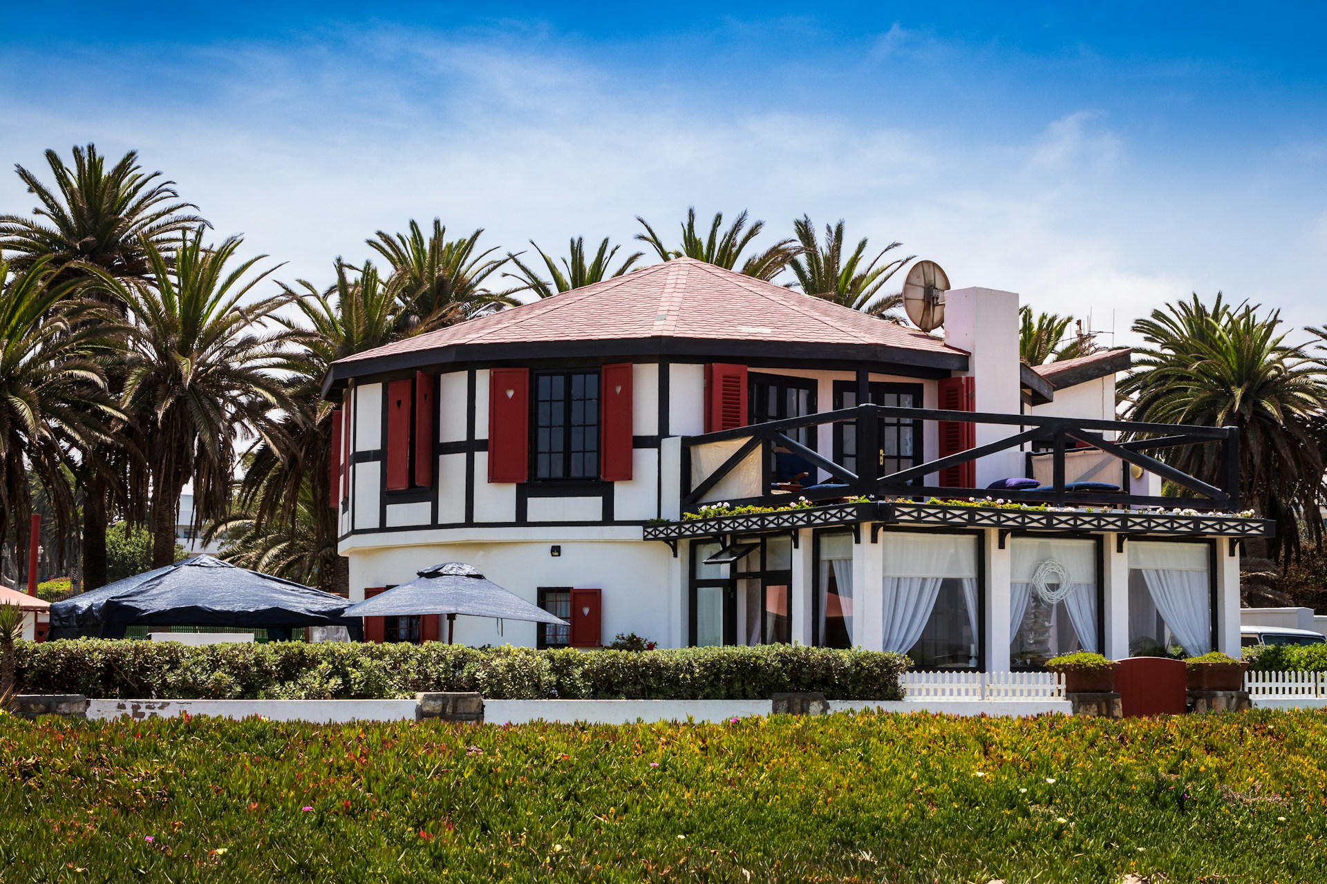 A two-story house with a black and white exterior and red accents is surrounded by lush greenery and palm trees. The house features large windows and a balcony on the upper floor, with a patio area below. The sky is clear and blue, suggesting a sunny day.
