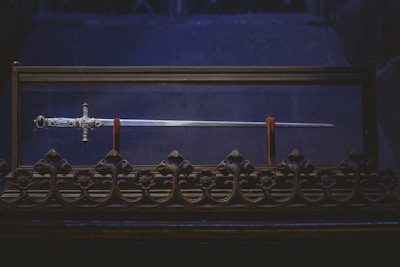 A beautifully crafted sword displayed in a glass case, featuring an intricately designed hilt and a long gleaming blade. The case rests on an ornate base with decorative floral patterns, set against a dark, ambient background.