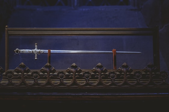 A beautifully crafted sword displayed in a glass case, featuring an intricately designed hilt and a long gleaming blade. The case rests on an ornate base with decorative floral patterns, set against a dark, ambient background.