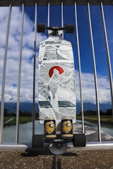 A longboard with intricate black and white artwork featuring geometric patterns and a red circular accent is propped against a metal railing. Below the deck, two cans of 'El Tony' mate are placed next to each other. The background includes a view of a river, green foliage, and a partly cloudy blue sky.