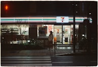 A convenience store is illuminated in the night, featuring bright signage and a recognizable logo. A person stands on the sidewalk under the neon light, with bicycles parked nearby. The scene includes a quiet street with a pedestrian crossing in front.