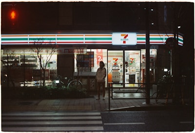 A convenience store is illuminated in the night, featuring bright signage and a recognizable logo. A person stands on the sidewalk under the neon light, with bicycles parked nearby. The scene includes a quiet street with a pedestrian crossing in front.