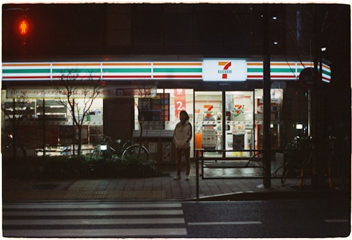 A convenience store is illuminated in the night, featuring bright signage and a recognizable logo. A person stands on the sidewalk under the neon light, with bicycles parked nearby. The scene includes a quiet street with a pedestrian crossing in front.