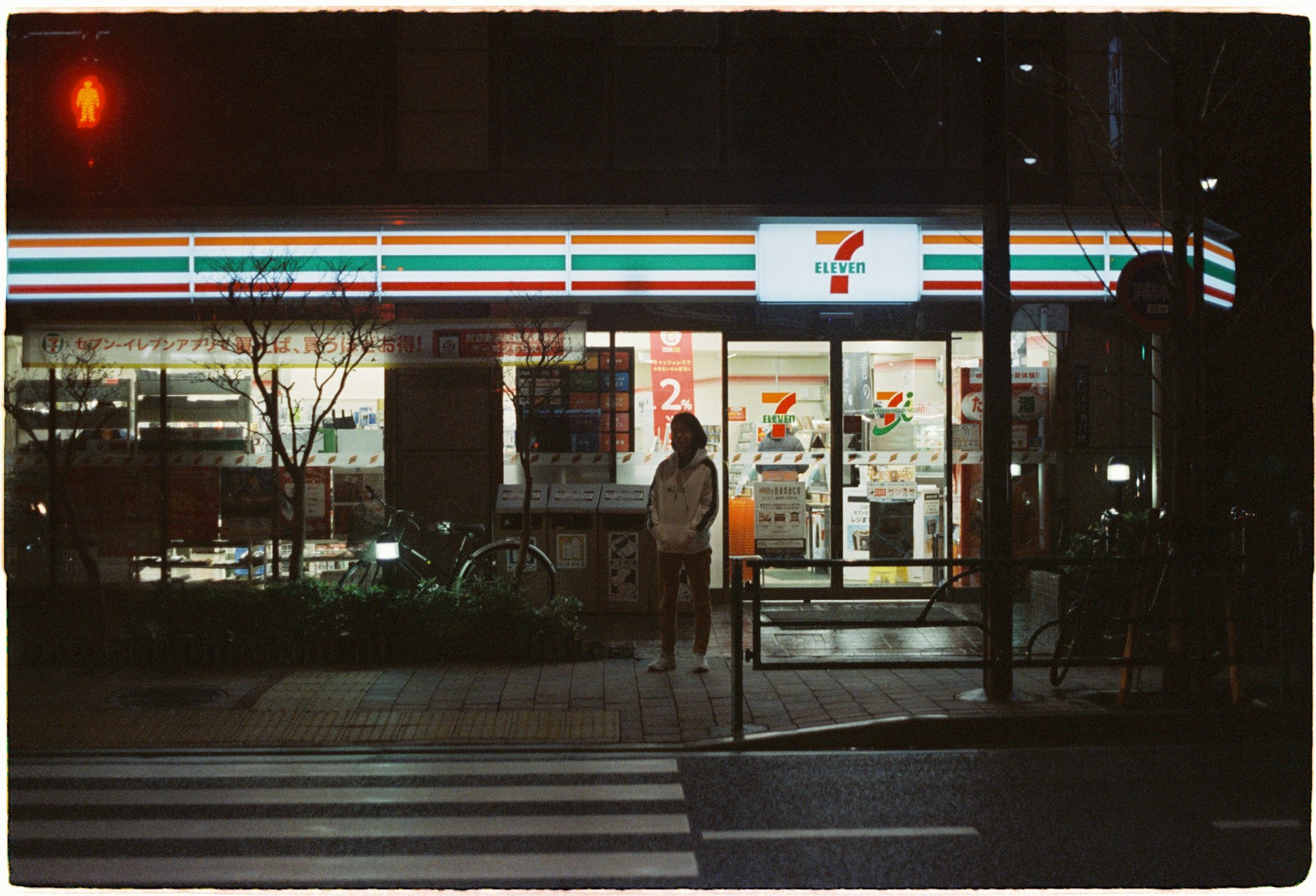 A convenience store is illuminated in the night, featuring bright signage and a recognizable logo. A person stands on the sidewalk under the neon light, with bicycles parked nearby. The scene includes a quiet street with a pedestrian crossing in front.