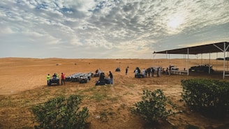 A desert landscape with a cloudy sky and a group of people gathered near all-terrain vehicles. Some people are wearing bright vests, and several vehicles are parked under a canopy. There is greenery in the foreground, and open sand dunes stretch into the distance.