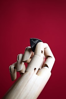 A wooden mannequin hand is gracefully holding a black guitar pick against a vivid red background. The fingers are articulated and showcase a lifelike pose, emphasizing the contrast between the natural wooden texture and the sleek, dark pick.