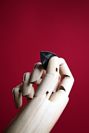 A wooden mannequin hand is gracefully holding a black guitar pick against a vivid red background. The fingers are articulated and showcase a lifelike pose, emphasizing the contrast between the natural wooden texture and the sleek, dark pick.
