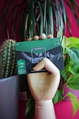 A wooden mannequin hand holds a green and black product package with text and graphics. The background features vibrant plants, including a cactus and large leafy green plant, set against a pink wall.