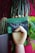 A wooden mannequin hand holds a green and black product package with text and graphics. The background features vibrant plants, including a cactus and large leafy green plant, set against a pink wall.