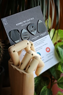 A wooden mannequin hand holds a black card featuring various guitar picks labeled as 'Rombo Try Out Mix Eco-Black'. The background includes blurred green plants and soft lighting.
