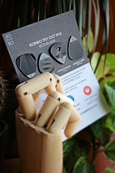 A wooden mannequin hand holds a black card featuring various guitar picks labeled as 'Rombo Try Out Mix Eco-Black'. The background includes blurred green plants and soft lighting.