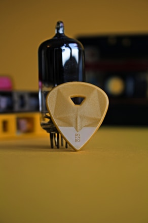 A close-up shot centered on a black vacuum tube and a yellow guitar pick with a triangular cutout near its top and the word 'ROBO' at the bottom. Both objects are placed on a surface with a yellow hue, and a blurred background with additional items in various colors can be seen.