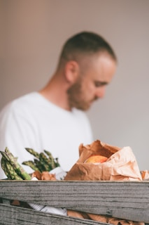 A person in a soft focus background is standing near a wooden crate containing fresh produce. The crate holds asparagus spears and a brown paper bag with a peach inside.