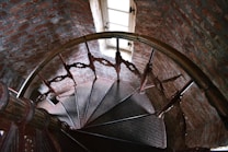 A spiral staircase made of dark wood and metal, with intricate balustrades, curves downward inside a narrow brick tower. A small window at the top allows natural light to enter, casting shadows on the textured bricks and the stairs.
