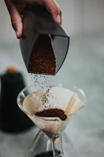 Close-up of a hand pouring freshly ground coffee into a simple brewing device on a wooden kitchen counter.