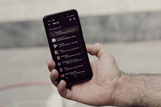 A person is holding a smartphone displaying a music playlist with various tracks listed. The screen shows song information with text and small album art thumbnails. The hand appears to be holding the phone firmly, and the background is blurred, focusing attention on the device.