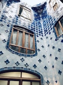 A building facade features a pattern of blue and white ceramic tiles with diamond-shaped accents. Wooden window frames are set within the tiled surface, creating an artistic blend of traditional and modern architecture.