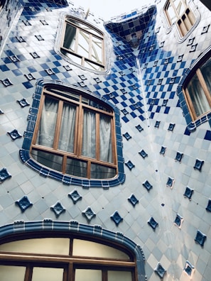 A building facade features a pattern of blue and white ceramic tiles with diamond-shaped accents. Wooden window frames are set within the tiled surface, creating an artistic blend of traditional and modern architecture.