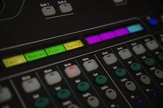 A close-up of an audio mixing console featuring various buttons and labeled controls. The panel includes labels like 'Muziek' and 'Film' with colored backlighting in green, yellow, and purple. Several rows of buttons are visible, each with different functions such as 'Mute' and 'Sel'. The overall look is technical and organized, suggesting a professional audio setup.