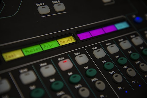 A close-up of an audio mixing console featuring various buttons and labeled controls. The panel includes labels like 'Muziek' and 'Film' with colored backlighting in green, yellow, and purple. Several rows of buttons are visible, each with different functions such as 'Mute' and 'Sel'. The overall look is technical and organized, suggesting a professional audio setup.