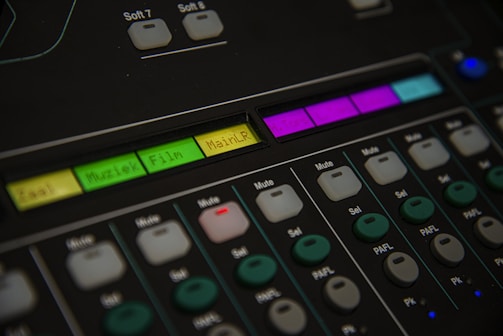 A close-up of an audio mixing console featuring various buttons and labeled controls. The panel includes labels like 'Muziek' and 'Film' with colored backlighting in green, yellow, and purple. Several rows of buttons are visible, each with different functions such as 'Mute' and 'Sel'. The overall look is technical and organized, suggesting a professional audio setup.