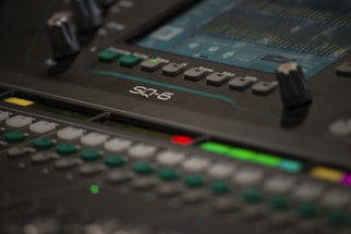 A close-up of a professional audio mixing console with various knobs, buttons, and a digital screen displaying sound levels. The screen shows multiple tracks and audio settings in detail.