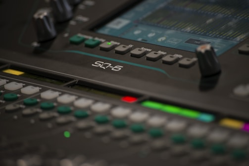 A close-up of a professional audio mixing console with various knobs, buttons, and a digital screen displaying sound levels. The screen shows multiple tracks and audio settings in detail.