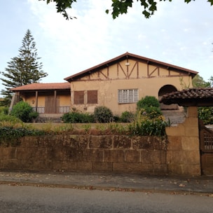 A rustic house with a beige facade and wooden accents is surrounded by greenery and shrubs. The roof is made of red tiles, and a large stone wall encloses the front yard. The entrance features steps leading to a wooden door, and a tall evergreen tree stands to the left of the house.
