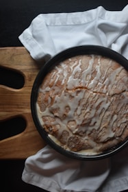 A round cake with a drizzle of white icing on top sits in a baking pan. The cake is placed on a wooden cutting board with a white cloth napkin partially draped around it.