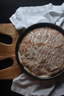 A round cake with a drizzle of white icing on top sits in a baking pan. The cake is placed on a wooden cutting board with a white cloth napkin partially draped around it.