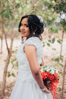 A bride in a white dress is standing amidst greenery, holding a bouquet of red and white flowers behind her back. Her hair is styled with a white floral accessory, and she is smiling slightly, looking over her shoulder.