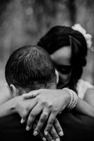 A black and white photograph captures an intimate embrace between two people. The focus is on their close contact, with a woman's hands resting gently on the man's shoulders, displaying a ring on her finger. Both subjects have their heads bowed slightly towards each other, suggesting a moment of tenderness and affection.