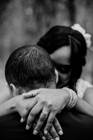 A black and white photograph captures an intimate embrace between two people. The focus is on their close contact, with a woman's hands resting gently on the man's shoulders, displaying a ring on her finger. Both subjects have their heads bowed slightly towards each other, suggesting a moment of tenderness and affection.