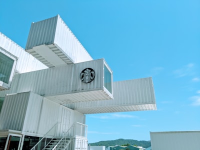 A modern building constructed from stacked shipping containers, adorned with the logo of a popular coffee chain. The containers are painted white and configured in an innovative design, standing out against a clear blue sky. A staircase and railing are visible, providing access between levels.