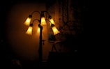 A multi-branched lamp with bell-shaped glass shades emits a warm, soft glow in a dimly lit room. The lamp's metallic structure is ornate, and the light creates gentle patterns on the surrounding surfaces.