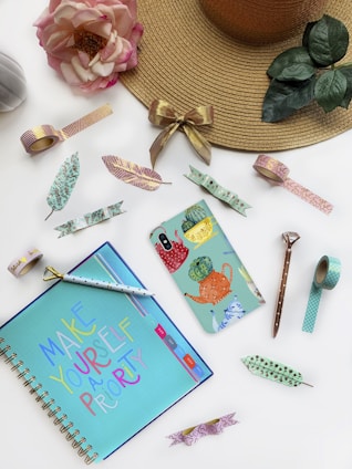 A decorative flat lay scene featuring a beige straw hat with a gold bow, a large pink rose, an open notebook with colorful text, and various patterned washi tapes and paperclips. A phone case with colorful cacti design is placed among them along with a stylish pen.