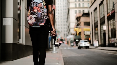 A person is walking down an urban street, carrying a backpack with Marvel superhero characters on it. They are holding a gimbal or stabilizer device, possibly indicating they are filming or vlogging. The street is lined with tall buildings, and a few cars are visible in the background, suggesting a busy city environment.