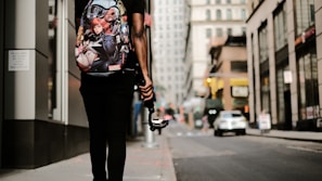 A person is walking down an urban street, carrying a backpack with Marvel superhero characters on it. They are holding a gimbal or stabilizer device, possibly indicating they are filming or vlogging. The street is lined with tall buildings, and a few cars are visible in the background, suggesting a busy city environment.