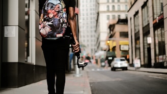 A person is walking down an urban street, carrying a backpack with Marvel superhero characters on it. They are holding a gimbal or stabilizer device, possibly indicating they are filming or vlogging. The street is lined with tall buildings, and a few cars are visible in the background, suggesting a busy city environment.