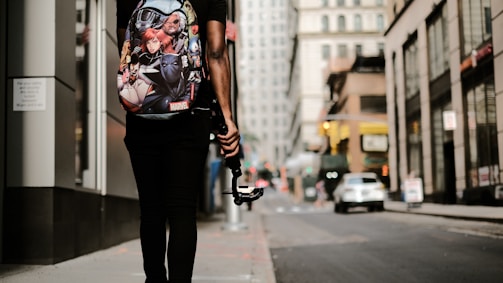 A person is walking down an urban street, carrying a backpack with Marvel superhero characters on it. They are holding a gimbal or stabilizer device, possibly indicating they are filming or vlogging. The street is lined with tall buildings, and a few cars are visible in the background, suggesting a busy city environment.