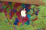 A large, white logo is prominently displayed against a vibrant, multicolored mural composed of flowing, abstract shapes. In the foreground, green foliage from trees partially obscures the view, adding a natural element to the lively background.