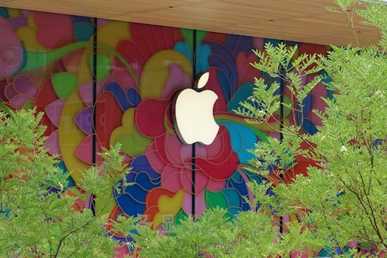 A large, white logo is prominently displayed against a vibrant, multicolored mural composed of flowing, abstract shapes. In the foreground, green foliage from trees partially obscures the view, adding a natural element to the lively background.