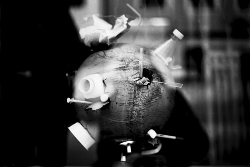 A monochrome image depicts a globe with various plastic items attached to it. These items include bottle caps, syringes, and other debris, suggesting themes of pollution and environmental impact.