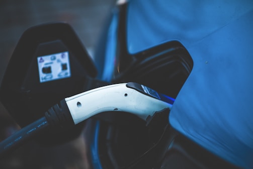 An electric vehicle charging plug is inserted into a charging port. The background features a vehicle with a blue surface, typically part of an electric car. A focus on modern technology and sustainable energy is evident.