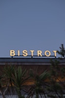 A sign with the word 'Bistrot' in bright, illuminated letters sits atop a building. Below, palm trees add a tropical feel to the scene. The background features an expansive sky, suggesting an outdoor setting and a calm atmosphere.