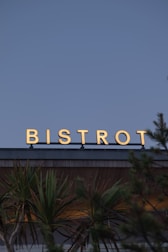 A sign with the word 'Bistrot' in bright, illuminated letters sits atop a building. Below, palm trees add a tropical feel to the scene. The background features an expansive sky, suggesting an outdoor setting and a calm atmosphere.