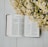 An open book featuring text in a non-Latin script is placed on a light wooden surface, accompanied by a bunch of small white and yellow flowers on the right side.